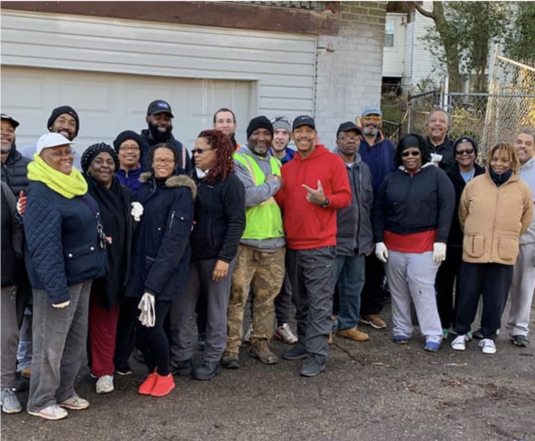 A group of 19 people in jackets and pants standing in front of a garage